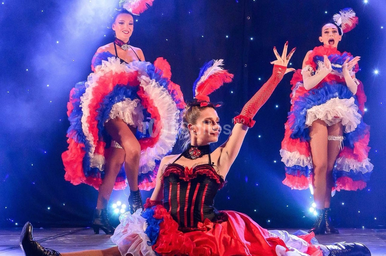 Danseuses de cabaret en costumes colorés rouge, blanc et bleu, exécutant une chorégraphie sur une scène éclairée.