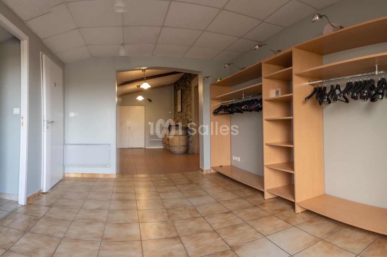 Vestiaire avec étagères en bois et portants à cintres, sol carrelé et vue sur un couloir menant à une pièce avec un tonneau.