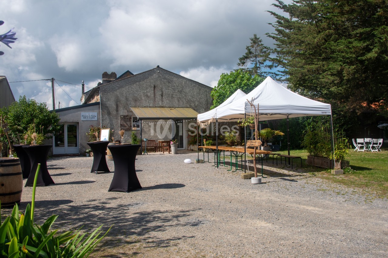 Cour extérieure avec tables, chaises et tonnelle blanche, devant un bâtiment en pierre entouré de végétation.