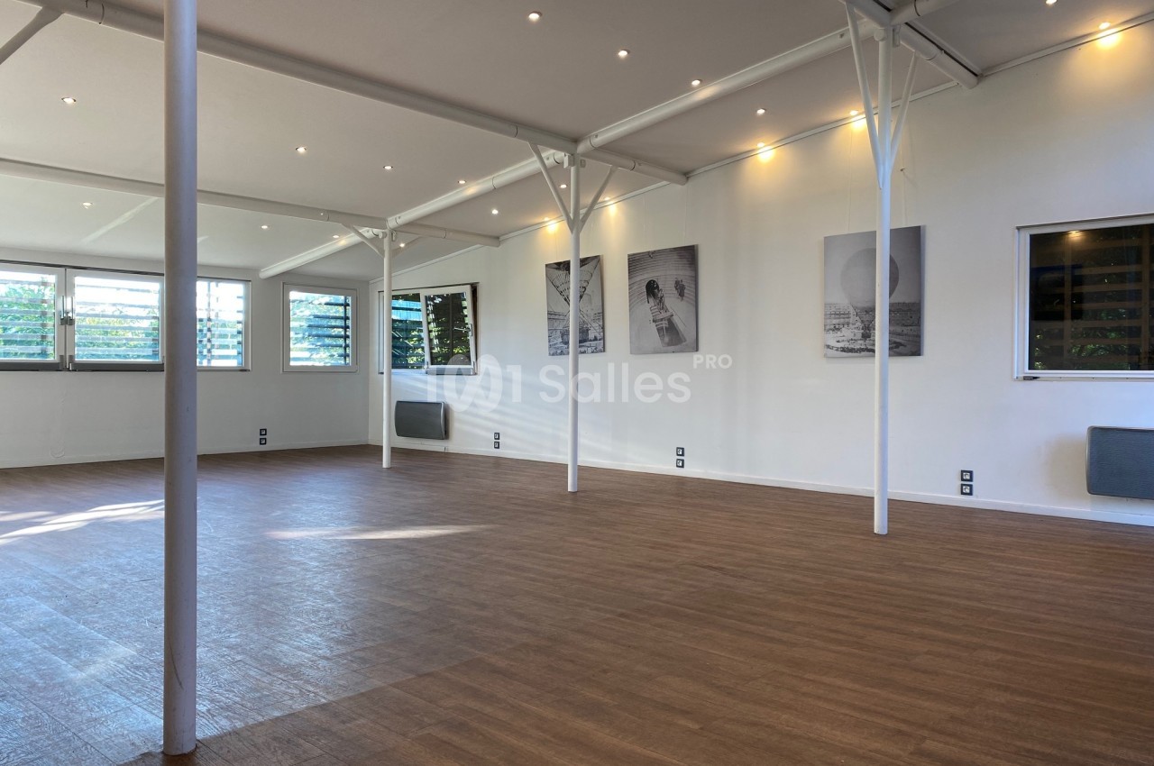 Salle lumineuse avec parquet en bois, murs blancs décorés de cadres, grandes fenêtres et éclairage encastré.