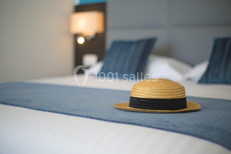 Chapeau de paille posé sur un lit avec une couverture bleue et des coussins assortis dans une chambre lumineuse.