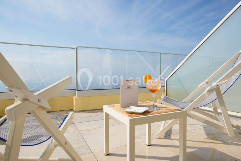 Deux chaises longues blanches sur une terrasse ensoleillée avec une table, un cocktail et une vue sur la mer.
