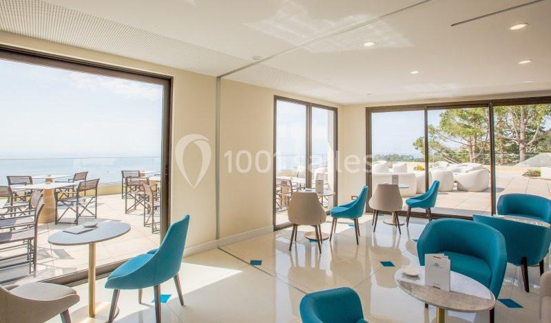 Salle lumineuse avec des chaises bleues et vue sur une terrasse aménagée donnant sur la mer.