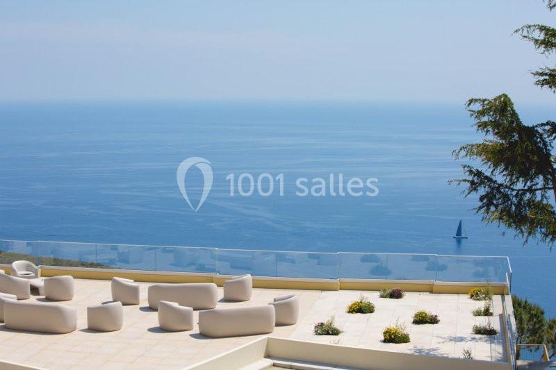 Terrasse avec mobilier moderne, vue sur la mer bleue et un voilier à l'horizon, entourée de végétation.