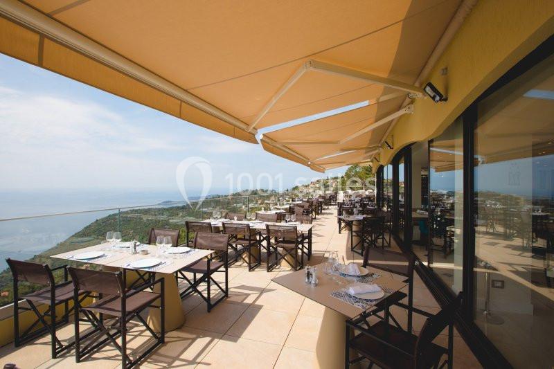 Terrasse d'un restaurant avec tables dressées, vue panoramique sur la mer et collines environnantes, sous des stores beige.