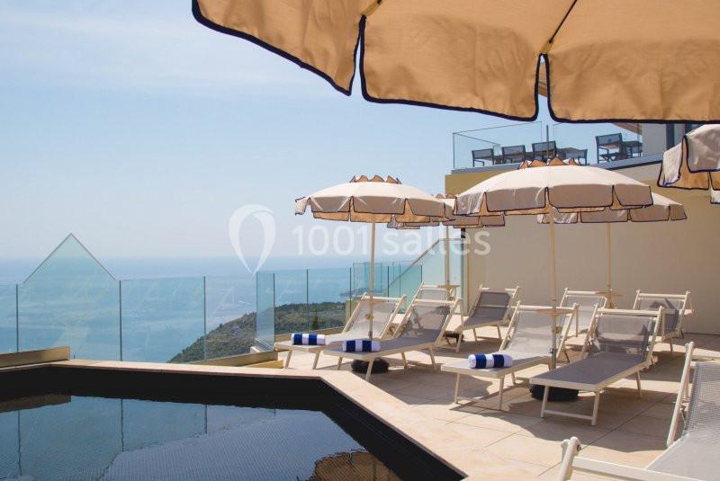 Terrasse avec transats, parasols beiges et vue sur la mer, entourée de balustrades en verre.