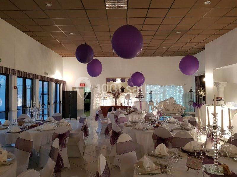 Salle de réception décorée avec des tables dressées, des chaises ornées de rubans violets et des ballons suspendus.