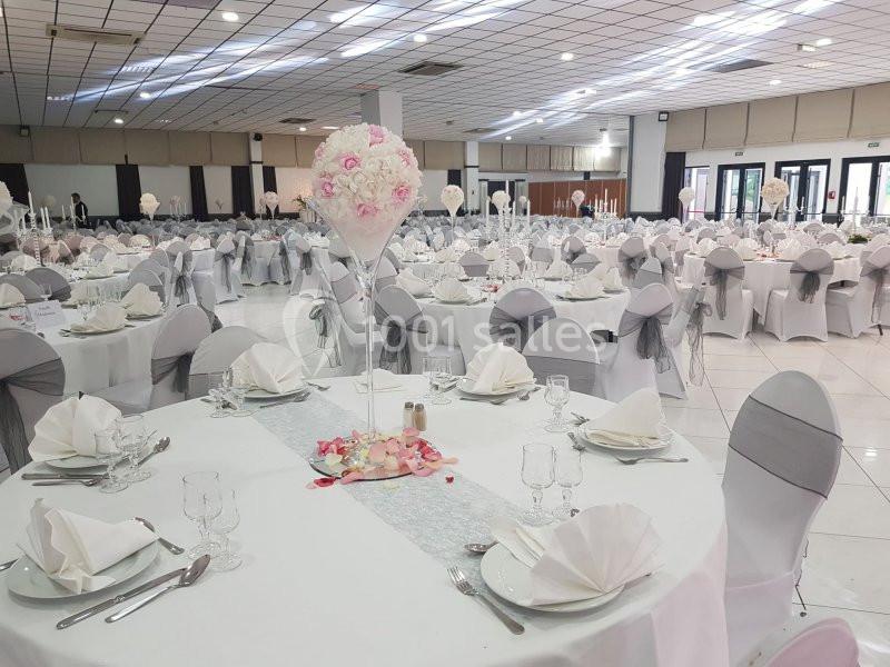 Salle de réception décorée avec des tables rondes, nappes blanches, chaises houssées et centre de table floral.