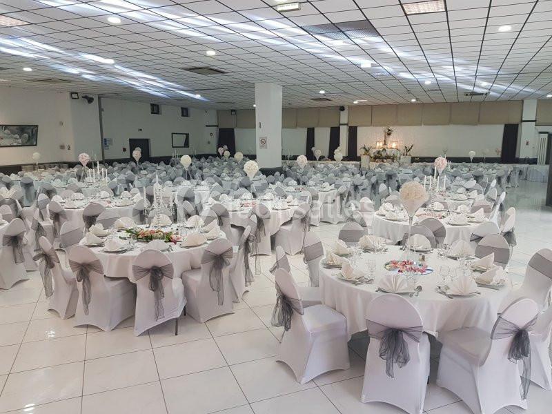 Salle de réception avec des tables rondes dressées, nappes blanches et chaises décorées de nœuds gris.