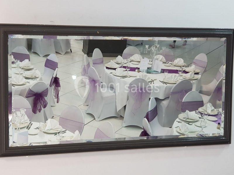 Salle de réception avec tables rondes dressées, chaises blanches décorées de nœuds violets, reflétées dans un miroir.