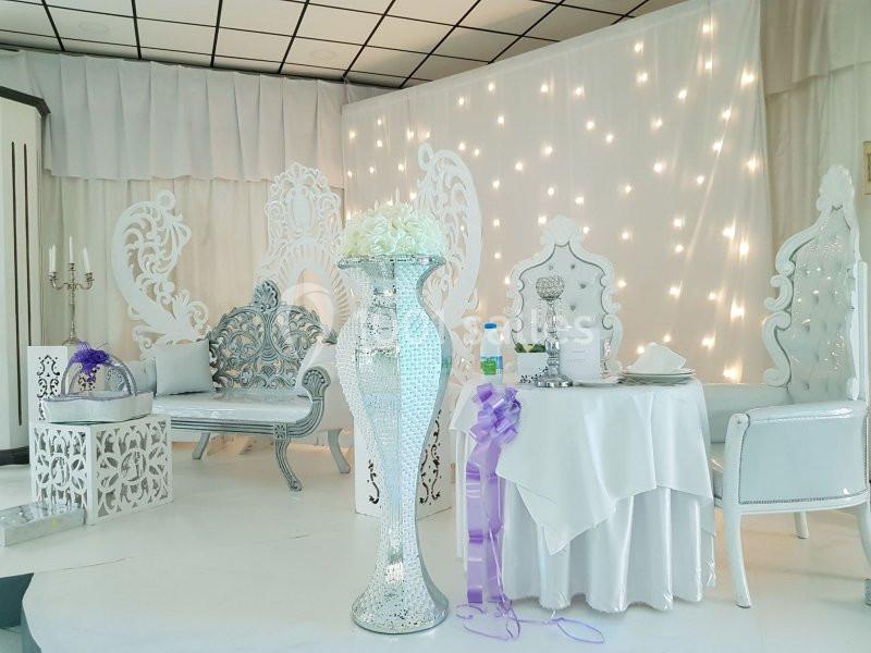 Décor de salle de réception avec mobilier blanc et argenté, vase central orné de fleurs et guirlandes lumineuses en arrière…