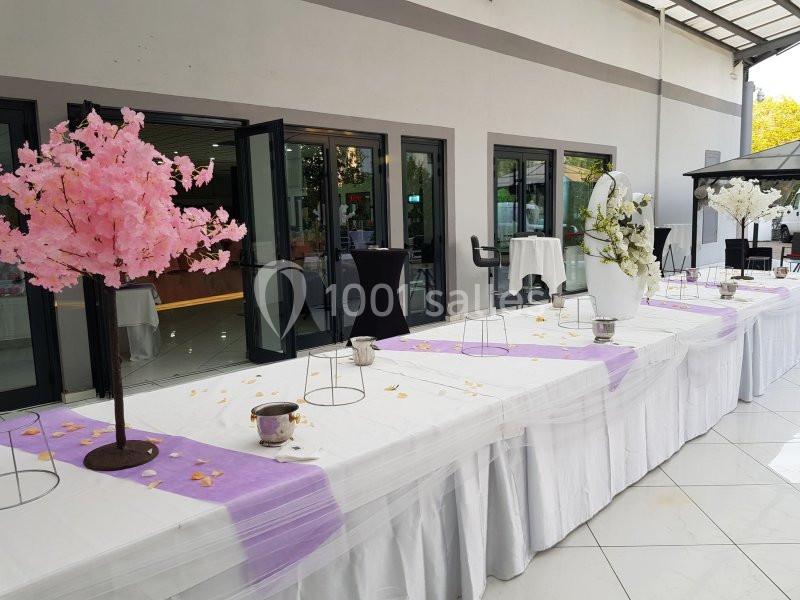 Table décorée pour un événement avec nappes blanches, chemins de table violets et arbres artificiels fleuris.