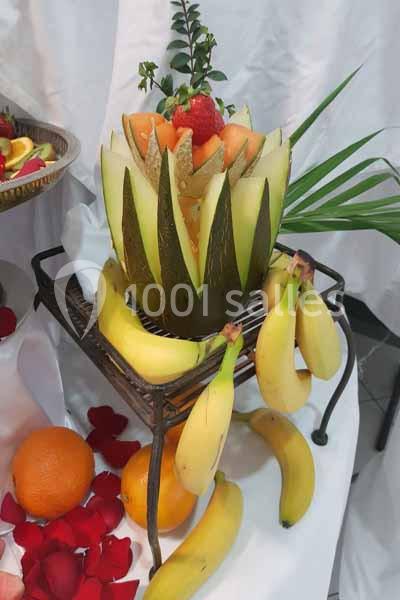 Melon sculpté rempli de fruits, entouré de bananes, oranges et pétales rouges, sur un support décoratif.