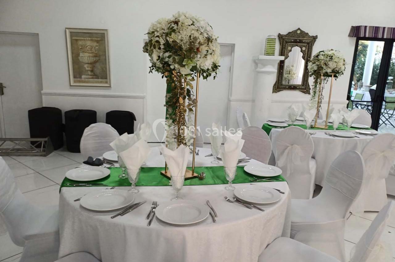 Salle de réception avec tables rondes dressées, nappes blanches, chemins de table verts et centres de table fleuris.