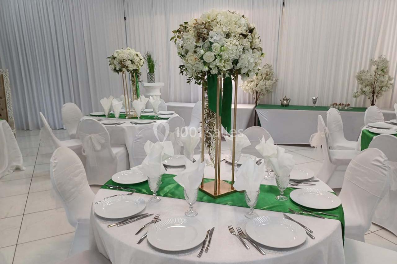 Salle de réception décorée avec des tables rondes dressées, nappes blanches, chemins de table verts et centres de table…