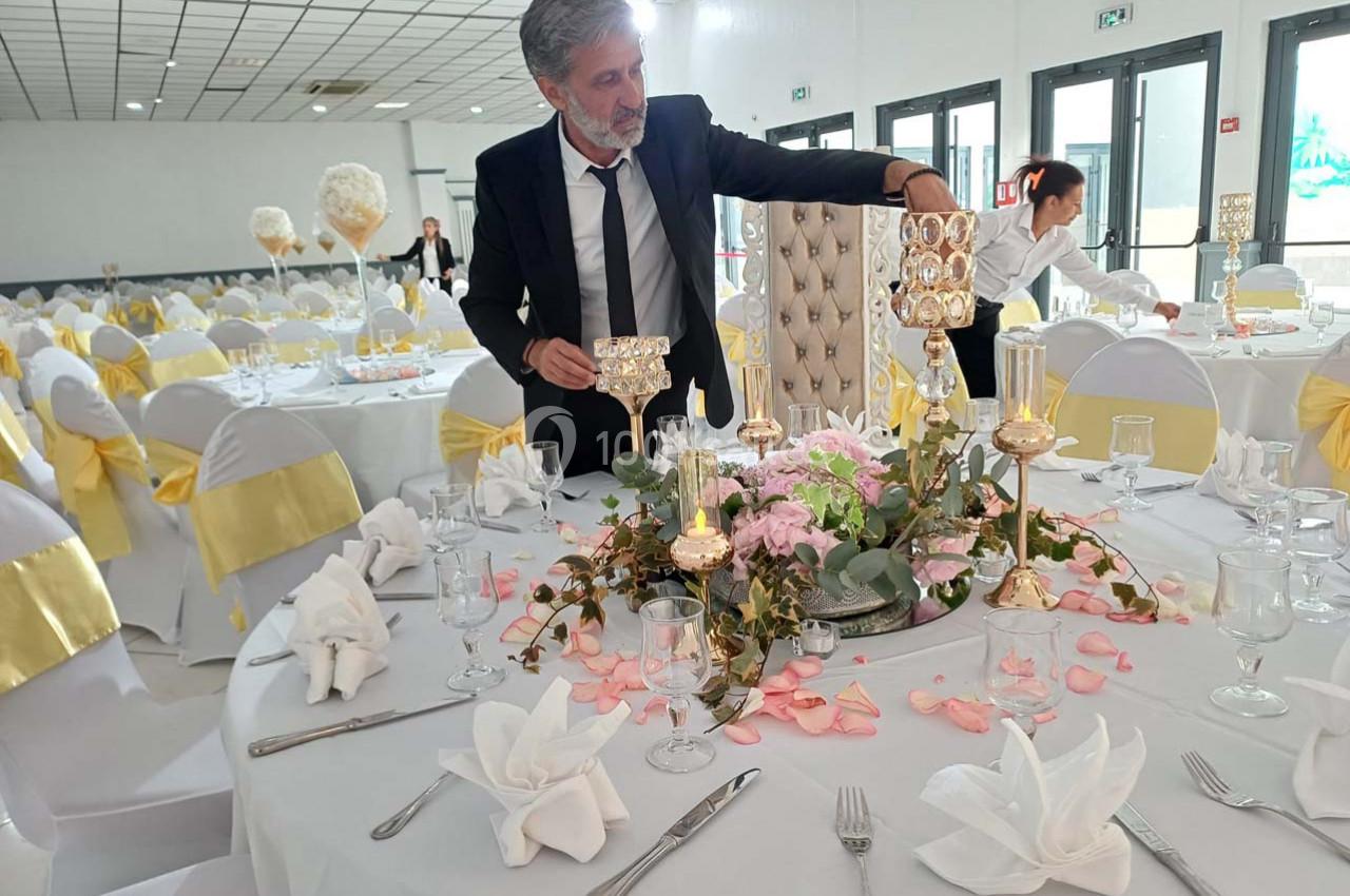 Un homme en costume ajuste la décoration florale et les bougies sur une table élégamment dressée dans une salle de réception.