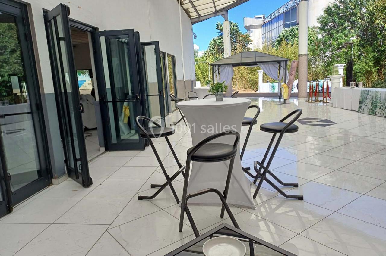 Terrasse lumineuse avec sol en carrelage blanc, table haute entourée de chaises et tonnelle visible en arrière-plan.