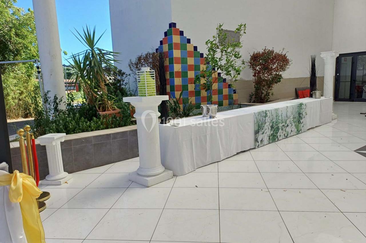 Table drapée de blanc avec colonnes décoratives, située dans un espace extérieur carrelé et entourée de verdure.