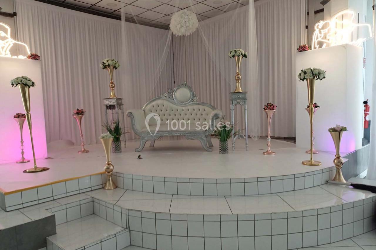 Décor de scène avec canapé argenté, colonnes dorées ornées de fleurs blanches et éclairage rose subtil.