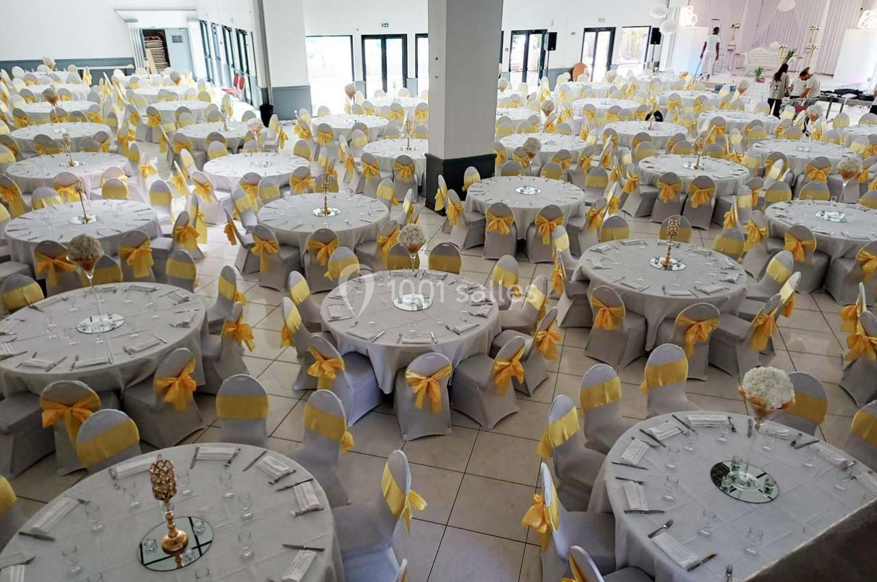 Salle de réception décorée avec des tables rondes, nappes blanches et chaises ornées de nœuds jaunes.