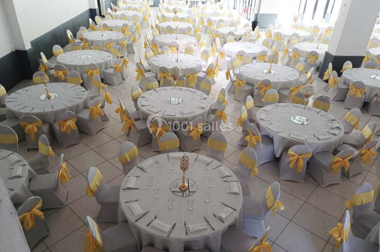 Salle de réception avec des tables rondes dressées, nappes blanches et chaises décorées de nœuds jaunes.