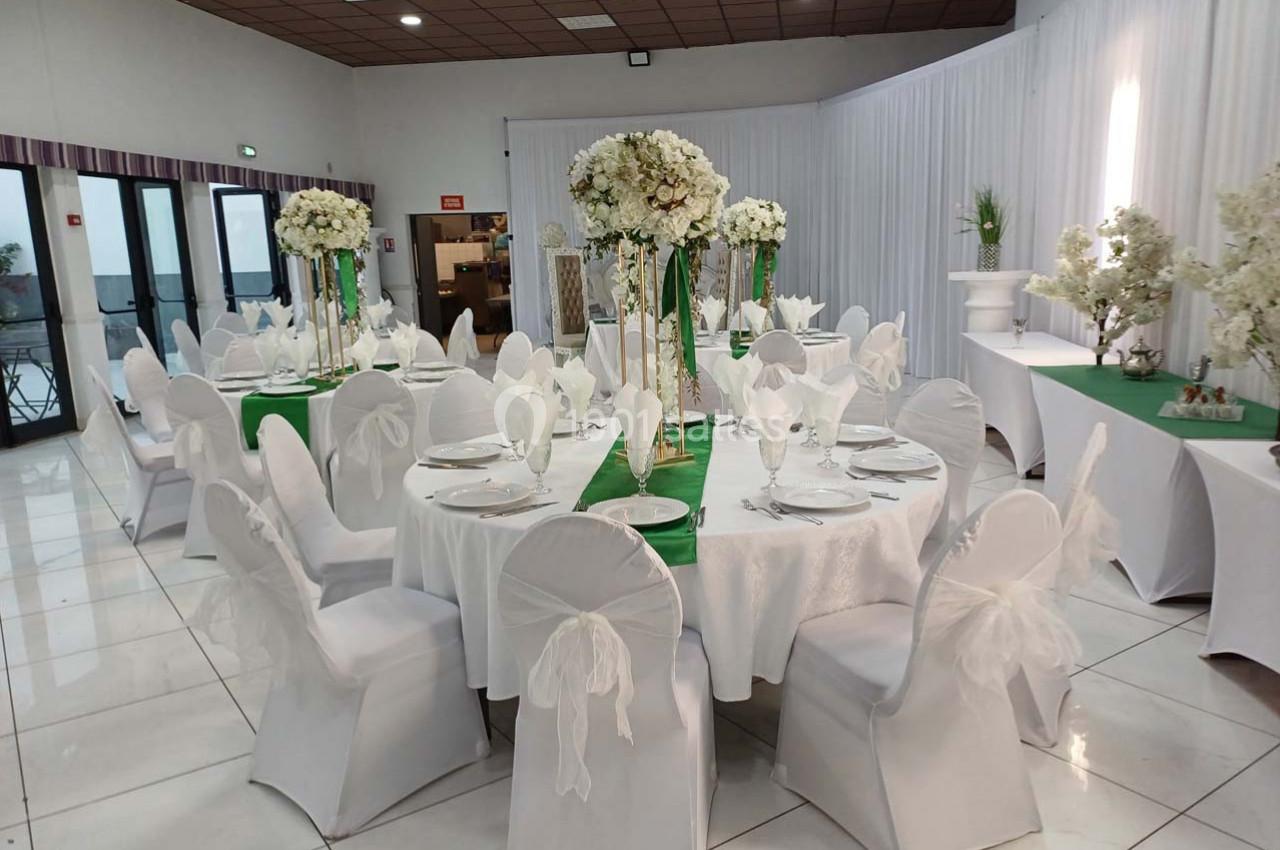 Salle de réception décorée avec des tables rondes, nappes blanches, chaises habillées et centres de table fleuris.