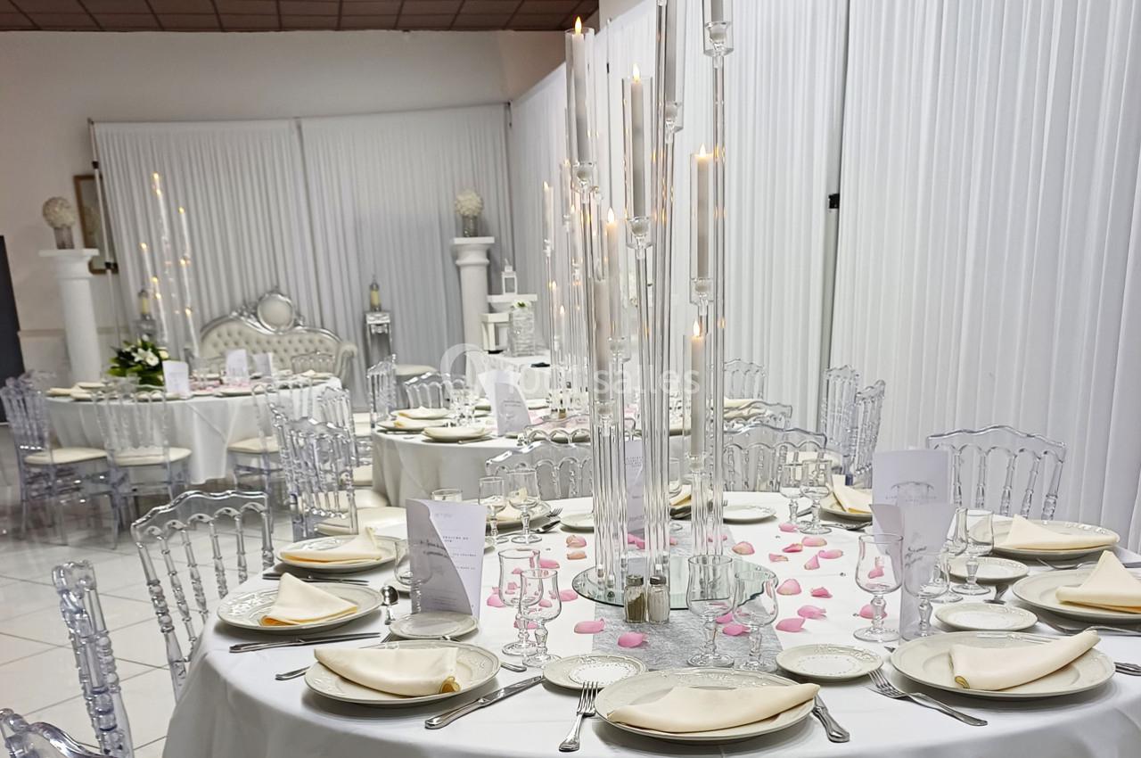 Salle de réception décorée avec des tables rondes, nappes blanches, chaises transparentes et centre de table lumineux.