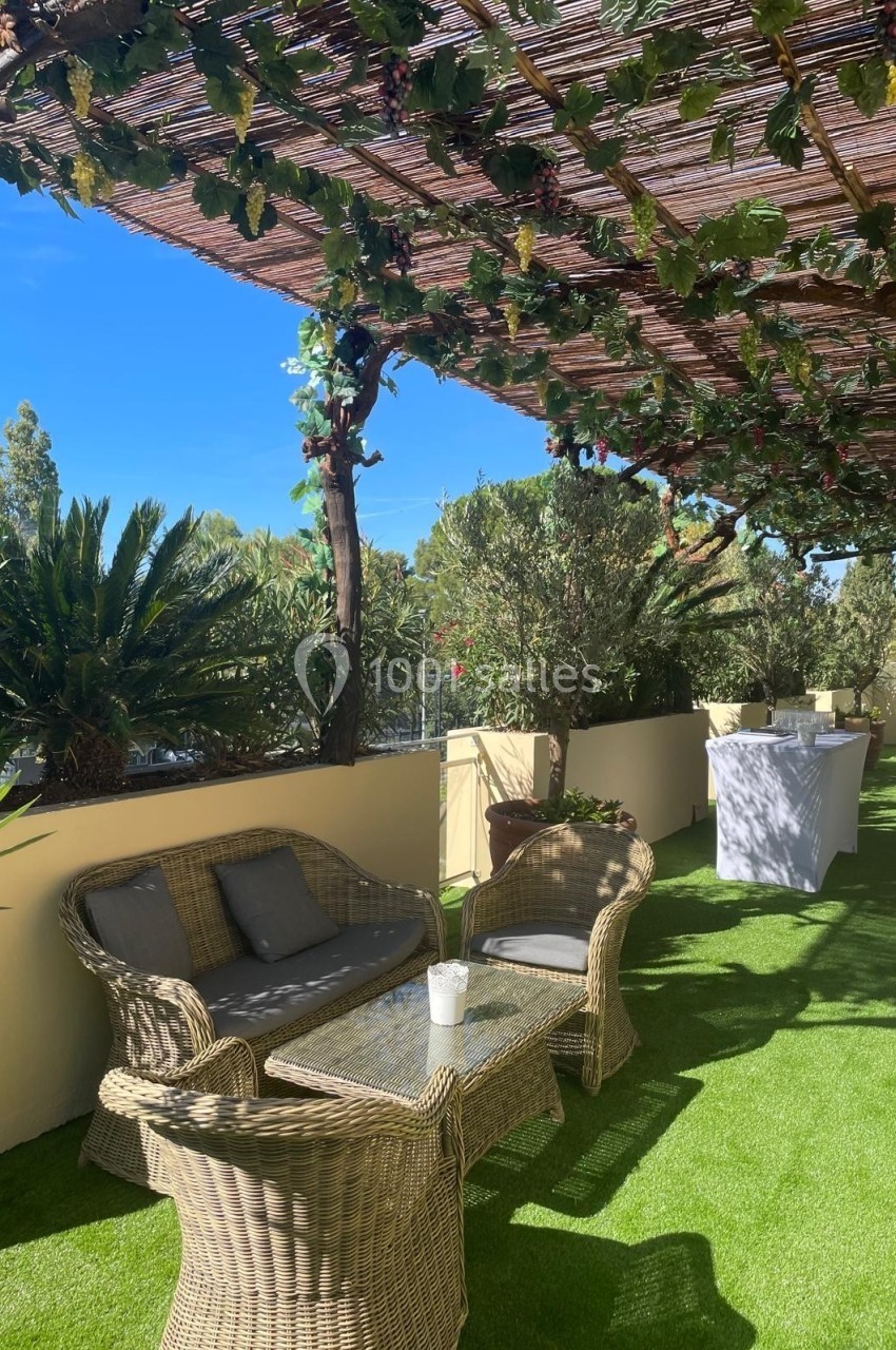 Terrasse avec fauteuils en rotin, table en verre, pergola couverte de feuillage et sol en gazon synthétique.
