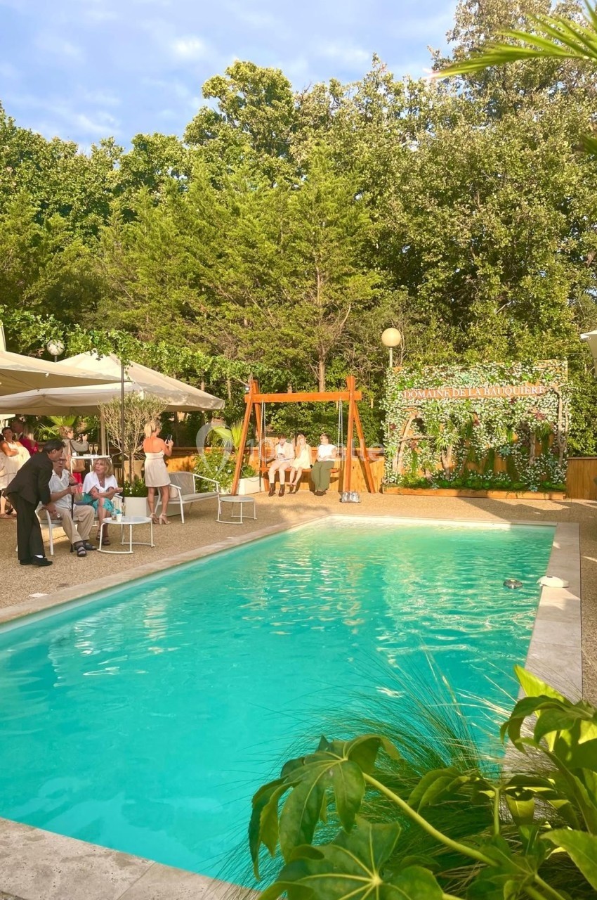 Piscine extérieure entourée de végétation, avec des personnes assises près d'une balançoire et d'un espace ombragé.