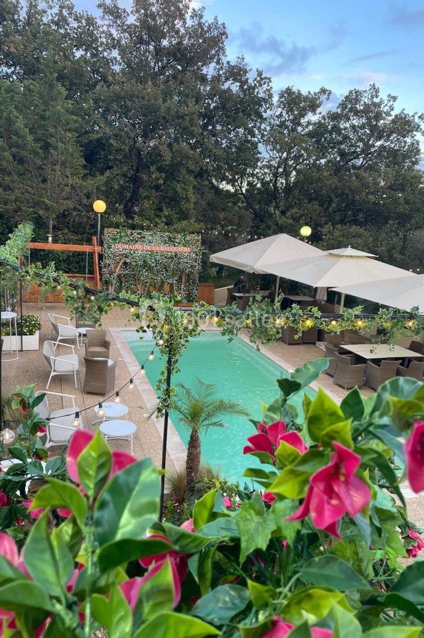 Piscine extérieure entourée de chaises, tables, parasols et végétation luxuriante sous un ciel partiellement nuageux.