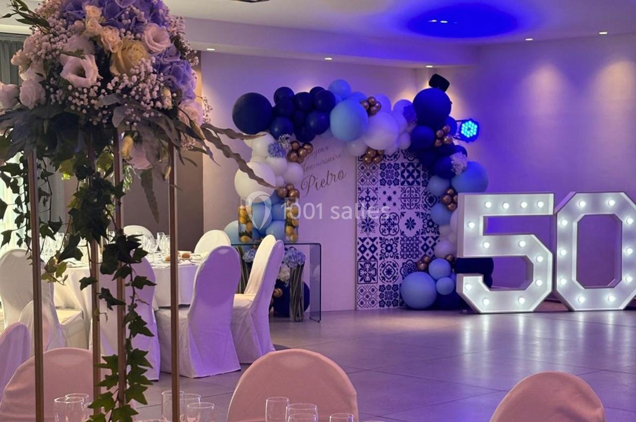 Salle décorée pour une fête avec ballons bleus et blancs, chiffre lumineux ’50’ et tables dressées avec fleurs.