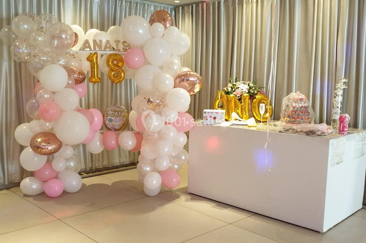 Décoration de fête avec arche de ballons roses et blancs, table ornée de fleurs, gâteaux et éléments festifs.