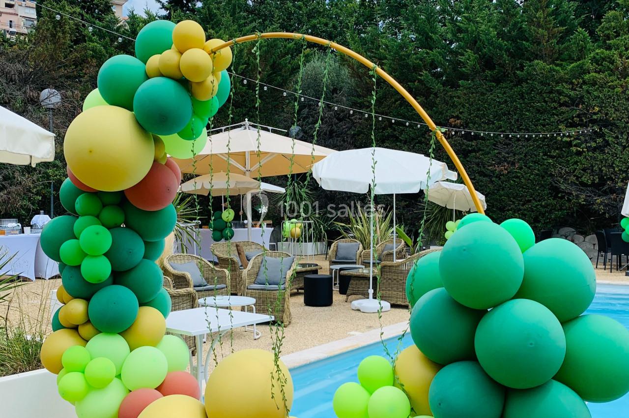 Arc décoratif en ballons colorés près d'une piscine, entouré de chaises et parasols dans un cadre extérieur verdoyant.