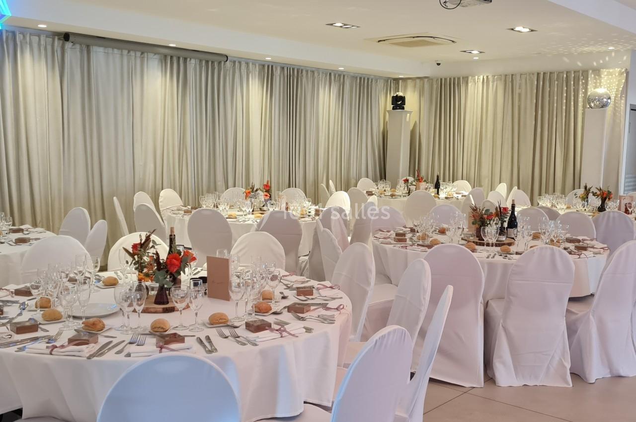 Salle de réception décorée avec des tables rondes dressées, nappes blanches et arrangements floraux rouges.