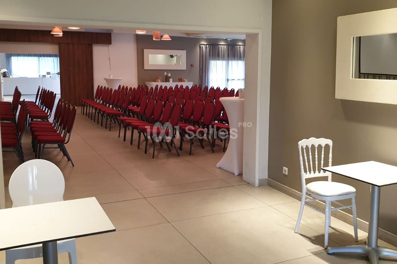 Salle avec des rangées de chaises rouges alignées, tables blanches au premier plan et éclairage naturel en arrière-plan.