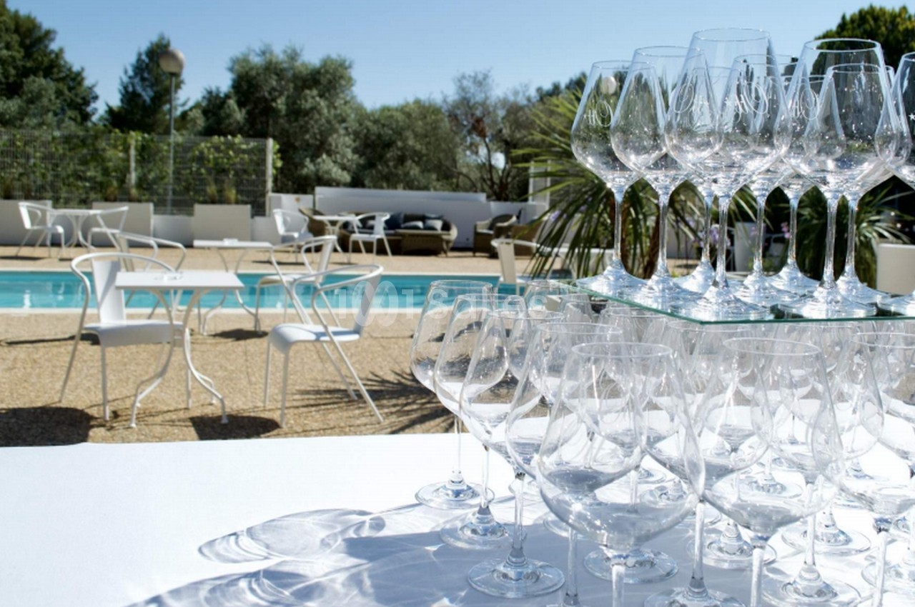 Verres à vin empilés sur une table blanche en extérieur, avec une piscine et des chaises en arrière-plan.