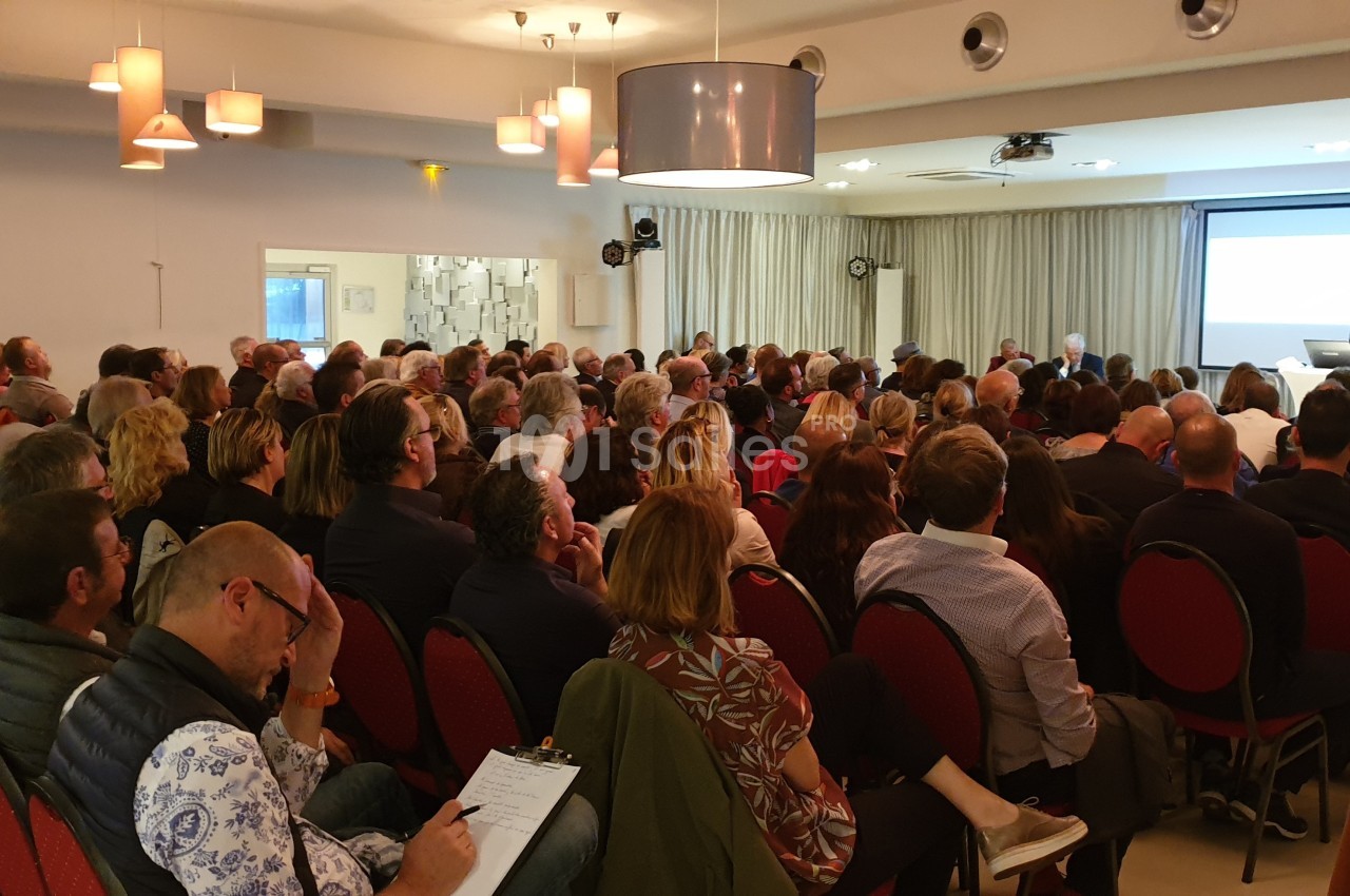 Un groupe de personnes assises dans une salle de conférence, écoutant un intervenant devant un écran de projection.
