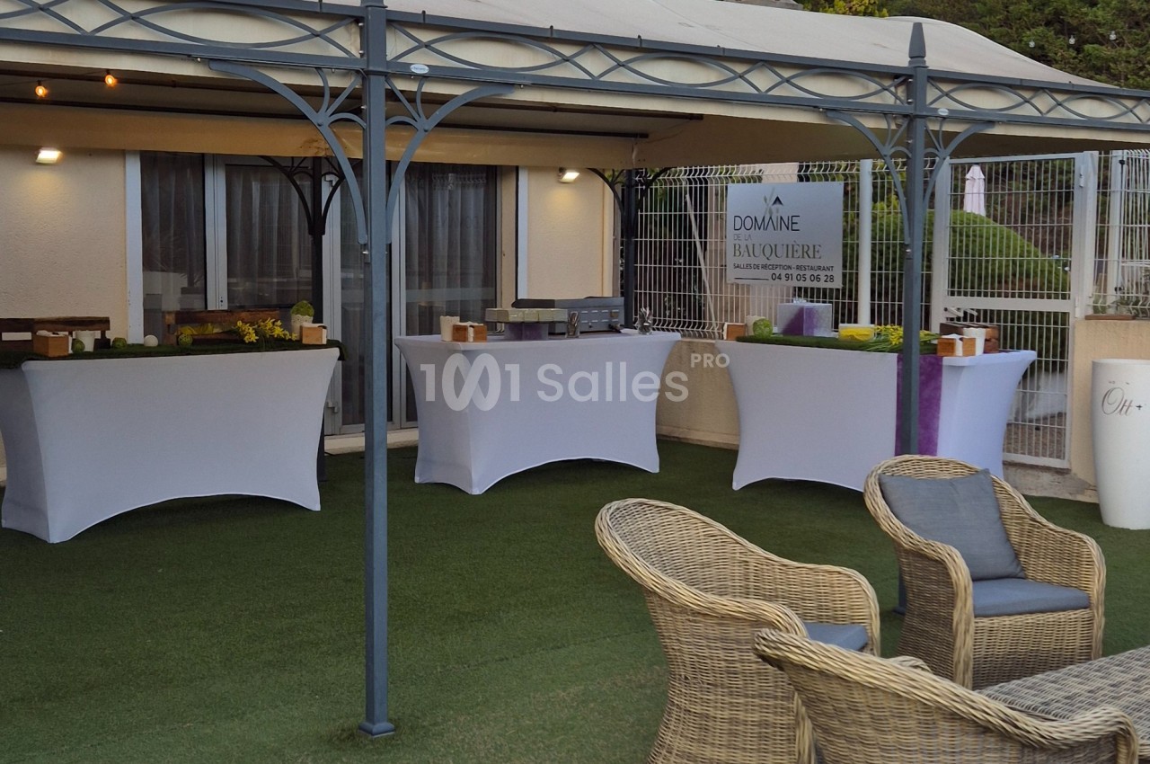 Tables habillées de nappes blanches sous une pergola, avec chaises en rotin au premier plan, dans un espace extérieur.