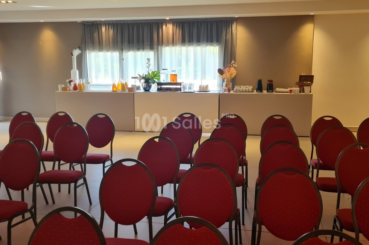 Salle de conférence avec des rangées de chaises rouges et une table de buffet en arrière-plan près de fenêtres.