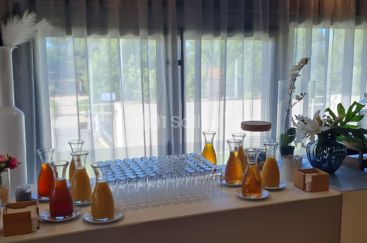 Buffet avec carafes de jus de fruits, verres alignés, décor floral et distributeurs de boissons devant une fenêtre.