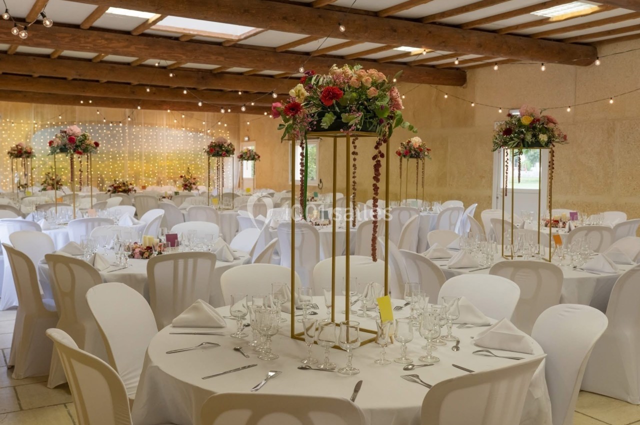 Salle de réception décorée avec des tables rondes, nappes blanches, centres de table fleuris et guirlandes lumineuses.