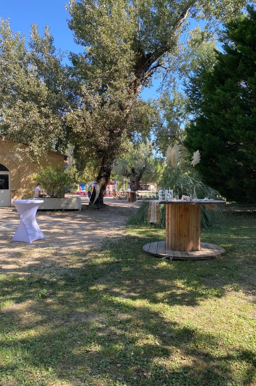 Table en bois décorée et table haute blanche installées dans un jardin ombragé avec des arbres et une pelouse.