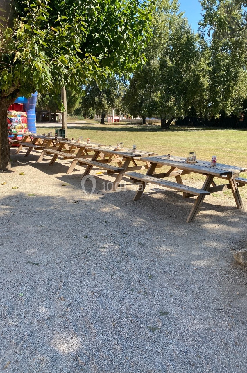Tables de pique-nique en bois alignées sous des arbres dans un espace extérieur ensoleillé avec des jeux gonflables en…