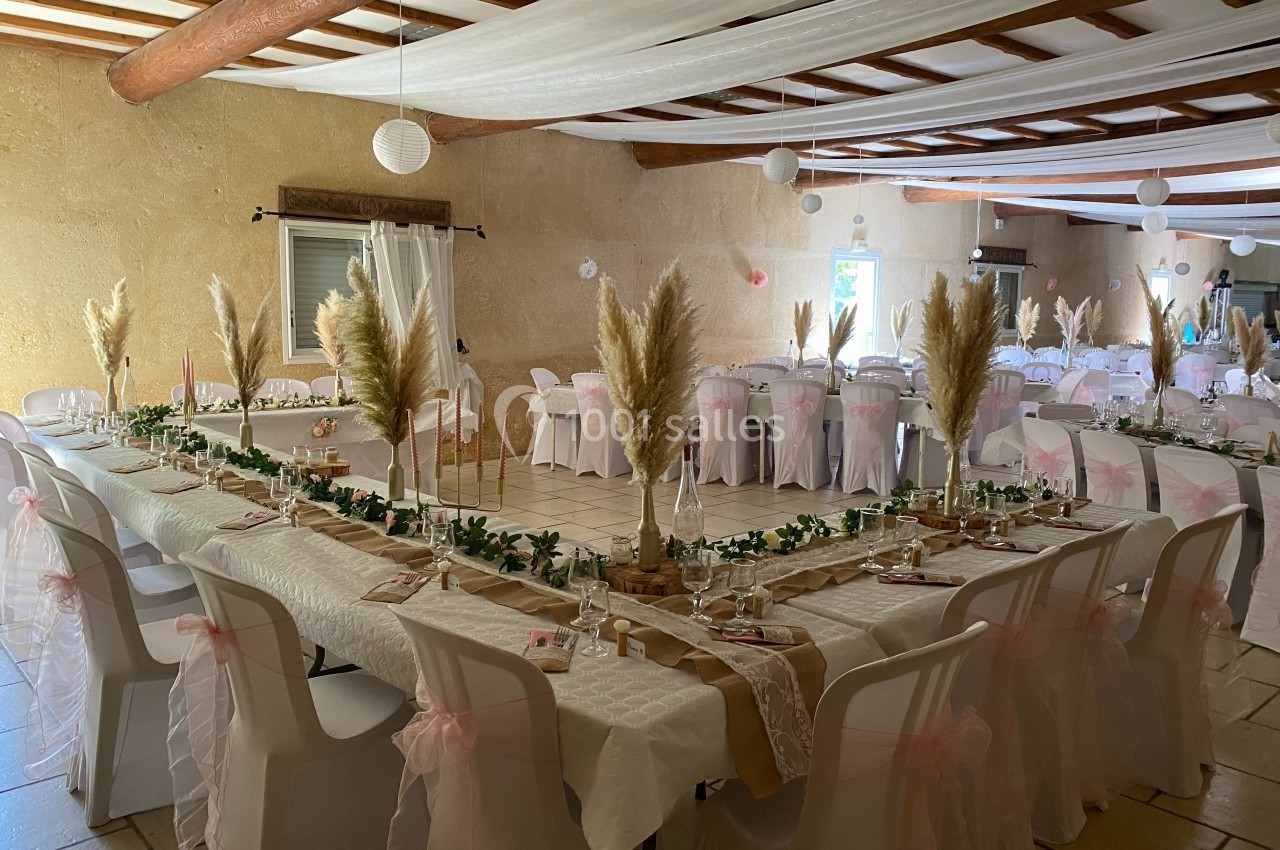 Salle de réception décorée avec des tables en U, nappes blanches, chaises ornées de rubans roses et ornements en pampas.