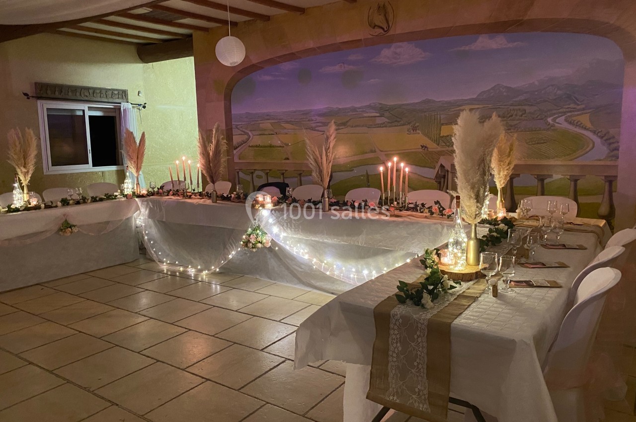 Salle décorée pour un événement, avec tables ornées de bougies, guirlandes lumineuses et pampas, devant une fresque murale.