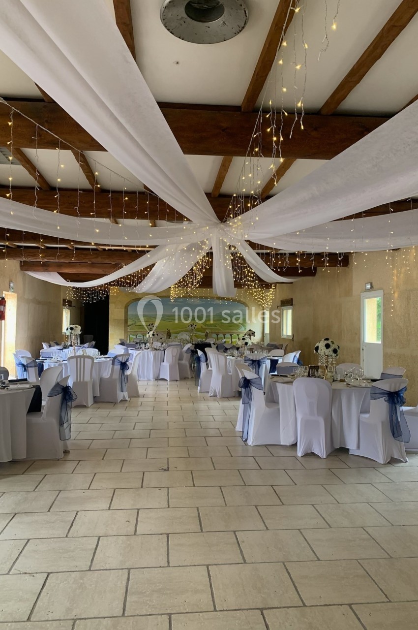 Salle de réception décorée avec des tables rondes, nappes blanches, nœuds bleus et guirlandes lumineuses suspendues.