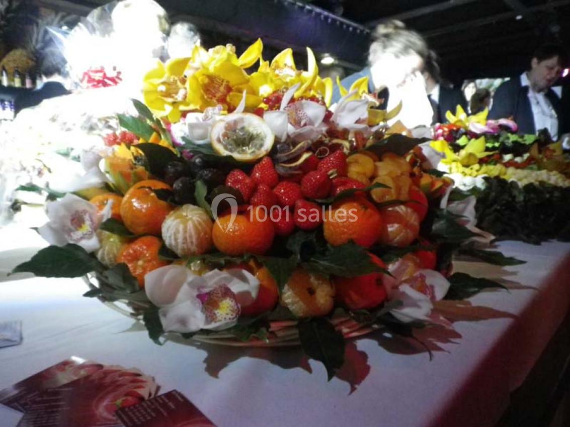 Corbeille garnie de fruits variés et fleurs décoratives, posée sur une table dans un environnement faiblement éclairé.