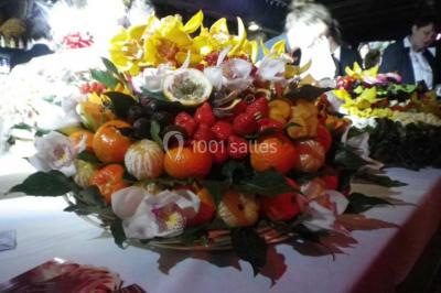 Corbeille garnie de fruits variés et fleurs décoratives, posée sur une table dans un environnement faiblement éclairé.