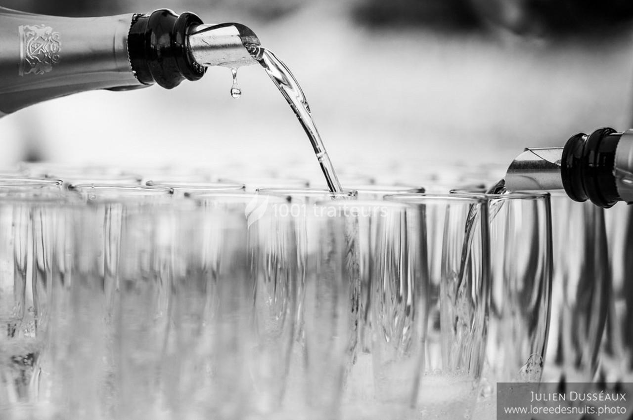 Deux bouteilles versent un liquide transparent dans des rangées de verres alignés, en noir et blanc.