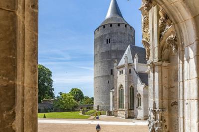 Un homme marche dans la cour d'un château médiéval avec une tour ronde et des bâtiments en pierre sous un ciel bleu.